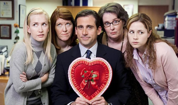 A man in a suit holds a red Valentine's heart while four women huddle around him with skeptical expressions.