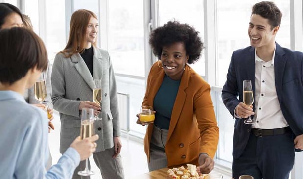 Young professionals at an office party, holding champagne glasses and snacks, smiling and chatting in a bright windowed space.