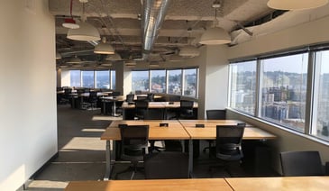 Modern open-plan office space with wooden desks, black ergonomic chairs, and floor-to-ceiling windows overlooking an urban cityscape.