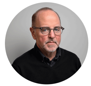 Portrait of Gregg Burke wearing glasses and a black sweater, posed against a soft light gray background.