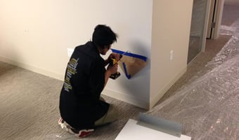 A person in a black shirt crouches while using a power tool on a wall, with protective floor coverings visible.