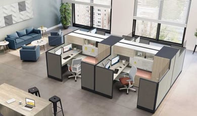 An open office layout featuring multiple workstations with grey dividers and a blue lounge area in the background.