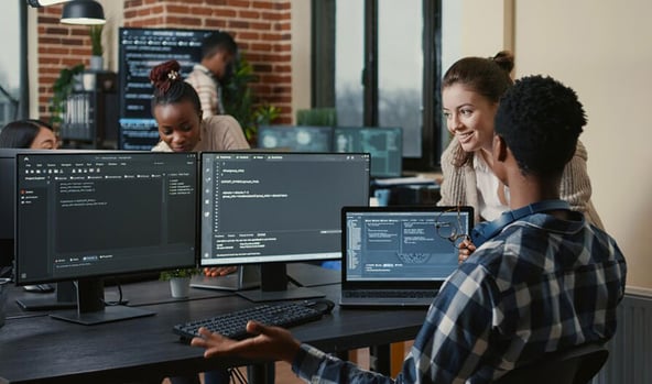 Programmers collaborating in a modern office, looking at code on multiple monitors while engaging in friendly discussion.