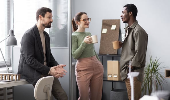 Three coworkers having a casual coffee break conversation in a modern office space.