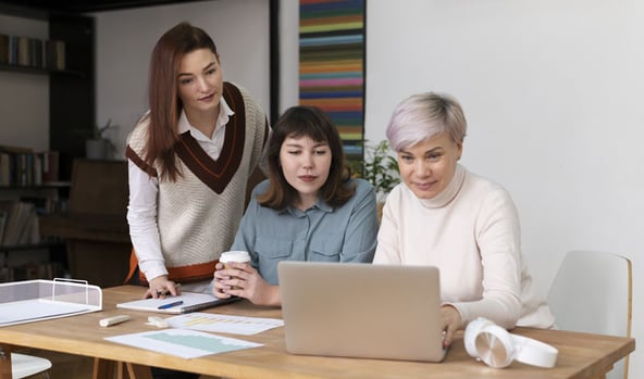 Three women collaborating around a laptop on a wooden desk in a cozy office setting.