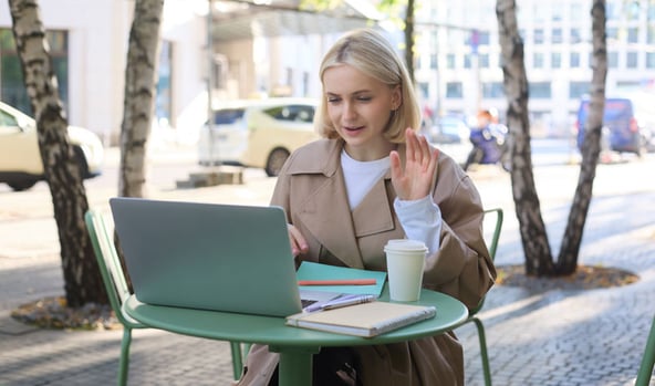 Young woman working on laptop at an outdoor café table, smiling while participating in a video call.