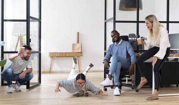 Office colleagues having fun during a break, with one person playfully riding a skateboard while others laugh and watch.