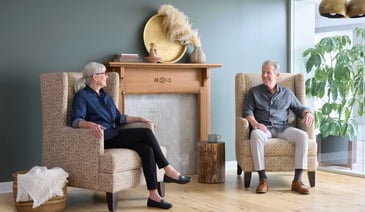 Two seniors relaxing in patterned armchairs by a fireplace in a warm, comfortable senior living lounge.