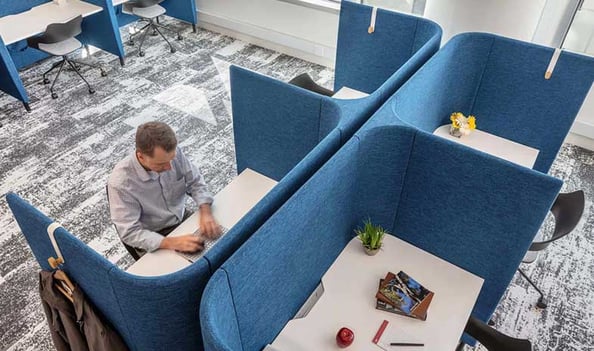 A worker using a private, blue upholstered workspace booth in a shared office environment.