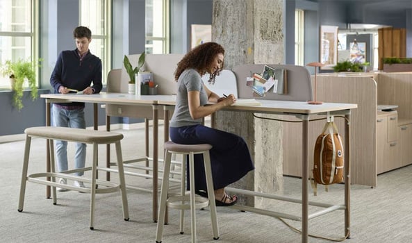 Two people working at a shared high desk in an open office with stools and personal storage.