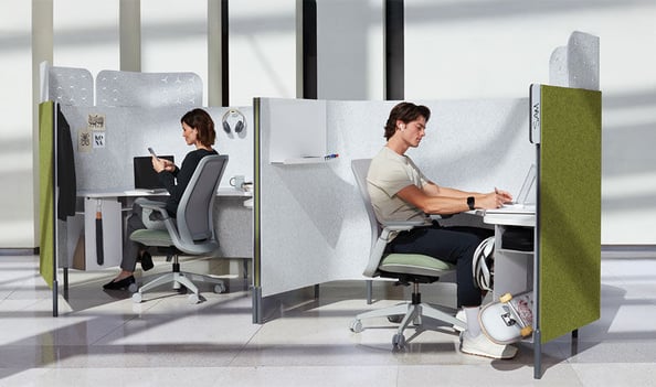 Two individuals seated in personal workspace pods, each with a partition for privacy and focus.