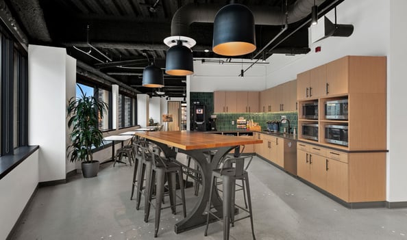 A contemporary office kitchen area with a large communal table, stools, and sleek lighting.