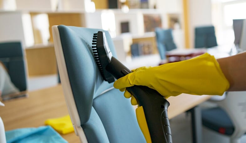 Person wearing yellow gloves cleaning an office chair with a vacuum brush attachment in a modern office setting.