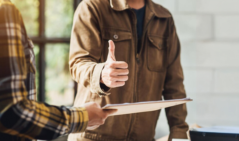 Two men standing. One man holds a clipboard with papers, while the other gives a thumbs-up gesture.