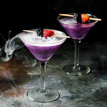 Two purple cocktails garnished with berries and dry ice creating a smoky effect.