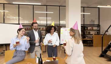 Four employees joyfully celebrate in an office space, donned in colorful party hats, while holding a cake and wine to mark a festive occasion.