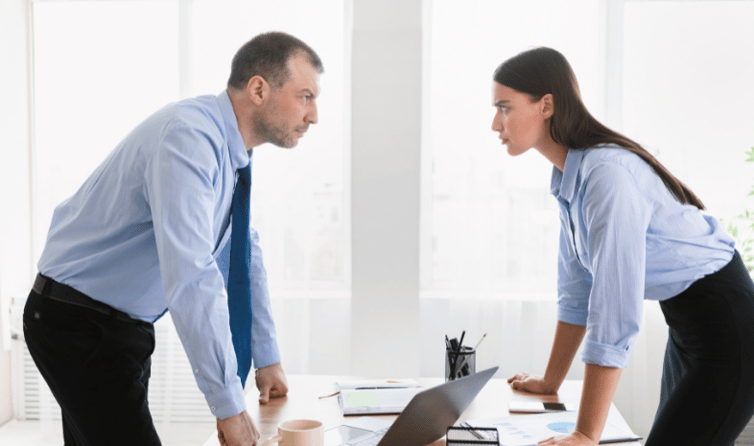 Two employees engaged in a heated discussion at their office desks, gesturing passionately while surrounded by paperwork and office supplies.