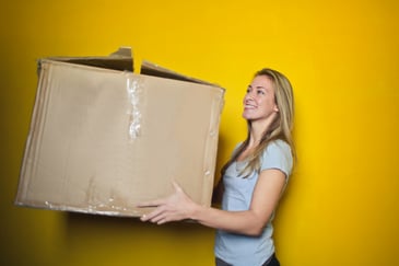 A woman in grey shirt holding a brown cardboard box.